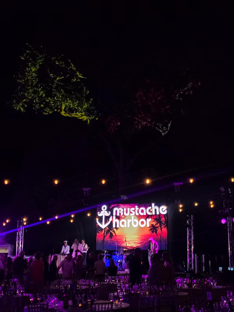 Live band on stage at a private outdoor gala, string lights and orange wash in the trees