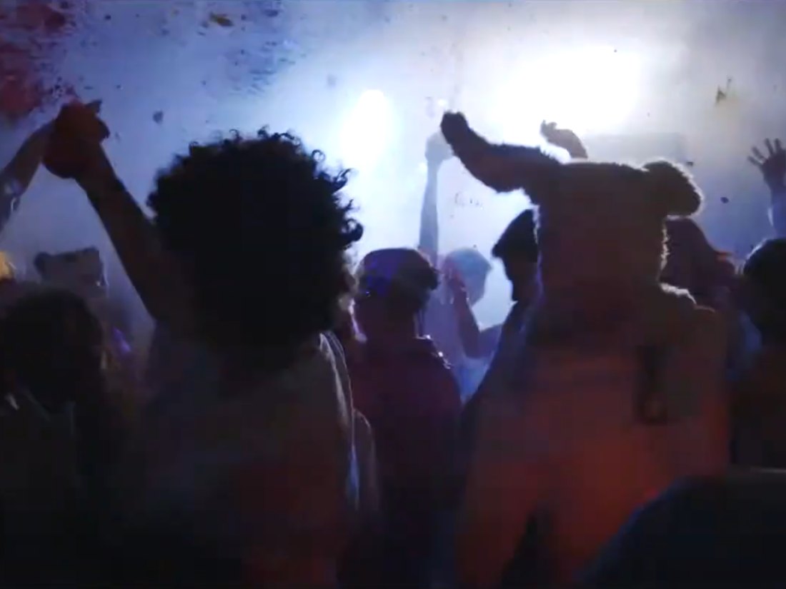 A packed warehouse dance floor with hands in the air, lit by stage lights through haze: a still from the 2016 short film The Cost of Color